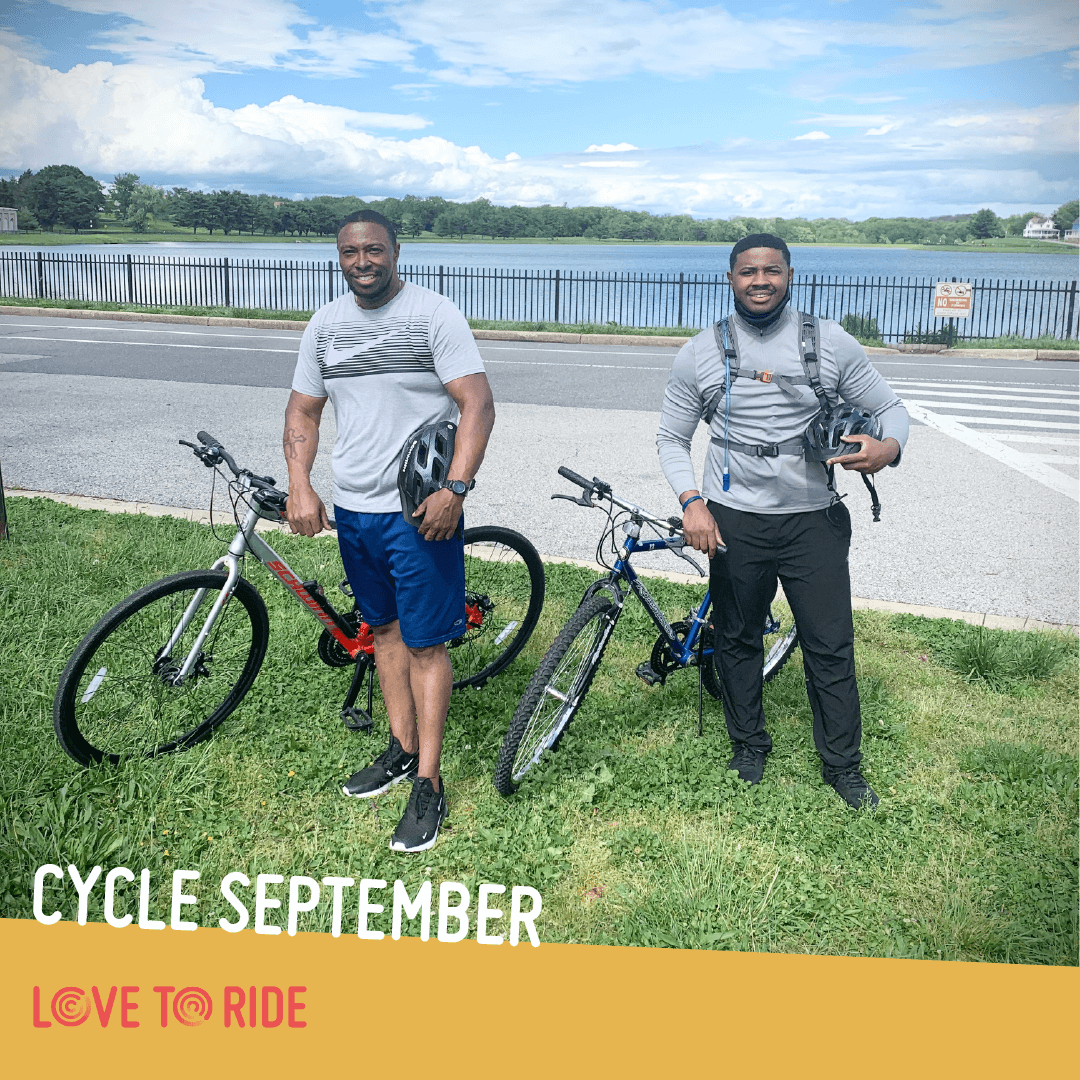 Two men stand with bikes on a grass verge next to a road and in front of a lake. They both hold helmets and are smiling. The text says ‘Cycle September’ and the Love to Ride logo is displayed. Two men stand with bikes on a grass verge next to a road and in front of a lake. They both hold helmets and are smiling. The text says ‘Cycle September’ and the Love to Ride logo is displayed.