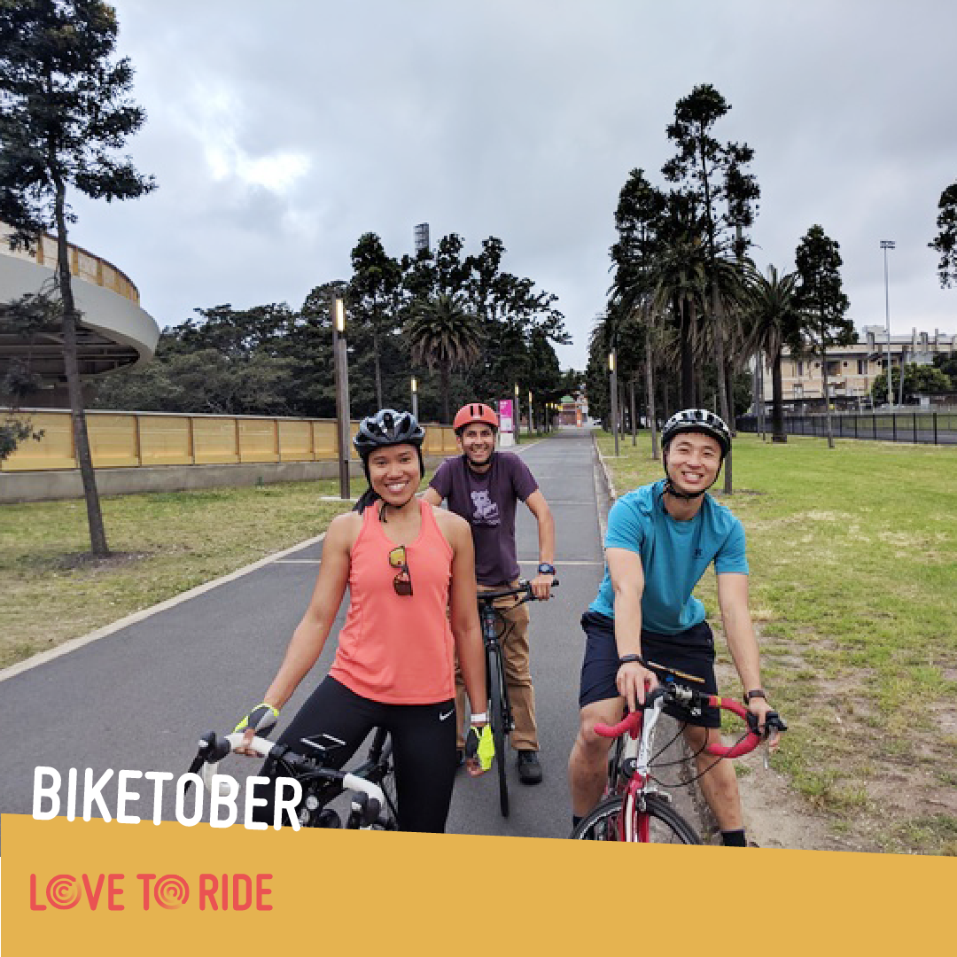 Three smiling riders pose on their bikes at the side of a paved bike path. In the background and along the edge of the path is grass, tall trees including Palm Trees, and some buildings. Three smiling riders pose on their bikes at the side of a paved bike path. In the background and along the edge of the path is grass, tall trees including Palm Trees, and some buildings.