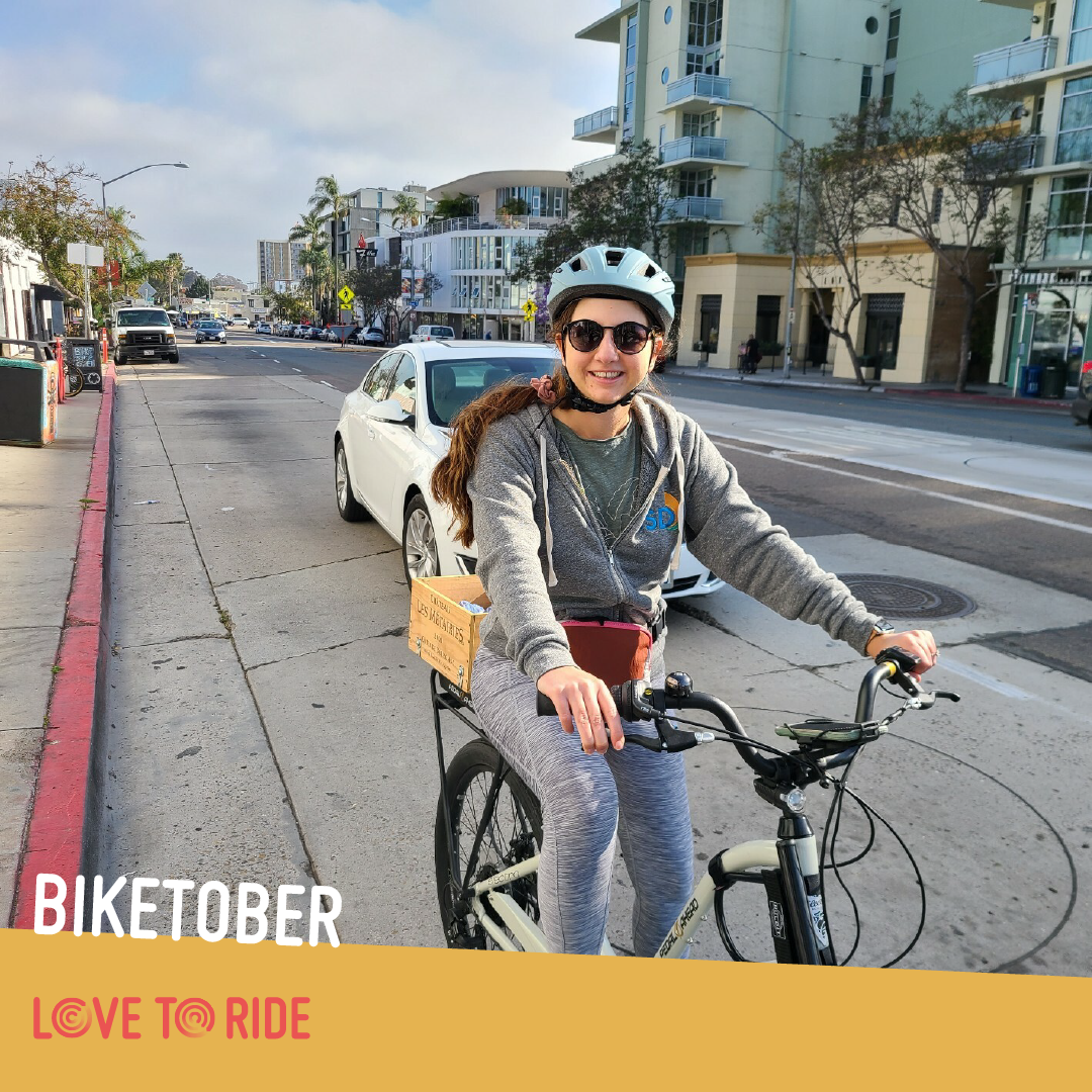 A woman on a bike smiles at the camera. She is at the side of an urban road and wearing a helmet and sunglasses. She rides a City Bike with a wooden box on the back. A woman on a bike smiles at the camera. She is at the side of an urban road and wearing a helmet and sunglasses. She rides a City Bike with a wooden box on the back.