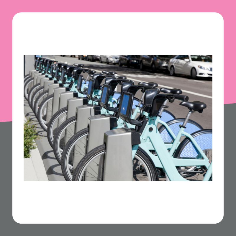 Image of hire bikes in a row on the street. The image has a pink and grey border. 