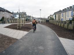 A person riding a bike on a smooth path which leads through a street of houses. The rider has a helmet and bag on their back and they are cycling away from the camera. 