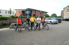 A group of people standing next to their bikes in a car park. There are 5 riders lined up next to eachother and there are buildings behind the car park. 