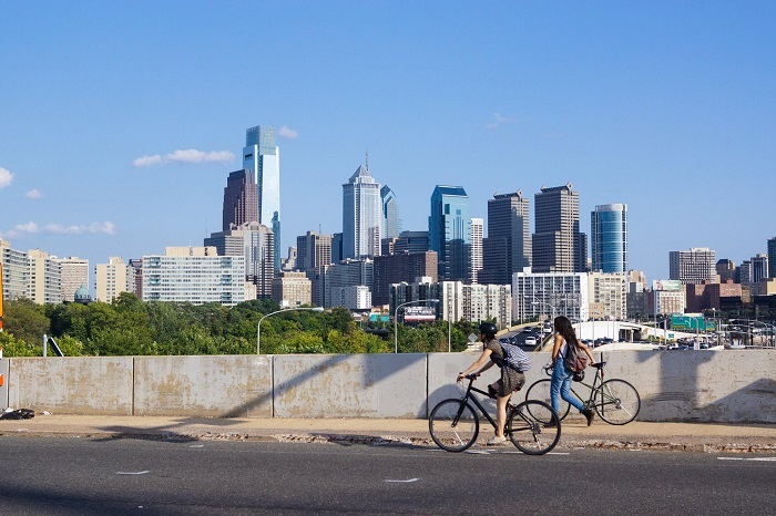 A person ride's their bike next to someone walking their bike with the Philly skyline behind them.